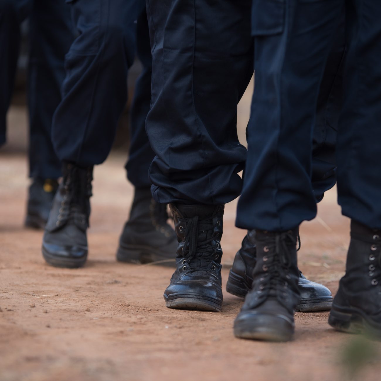 A group of people in dark Bamogale Security uniforms and black boots walk together on a dirt path, only their lower legs and feet visible.